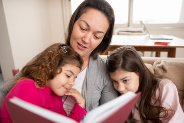 Lovely latin mother sitting in sofa reading for her kids. Indoors at home living room. Togetherness, multi-generation family, support concept.