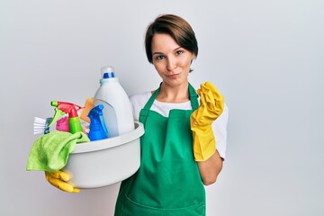 Young brunette woman with short hair wearing apron holding cleaning products doing money gesture...
