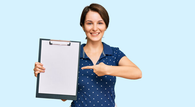 Young brunette woman with short hair holding clipboard with blank space smiling happy pointing with hand and finger
