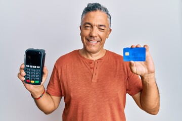 Handsome middle age mature man holding dataphone and credit card winking looking at the camera with...