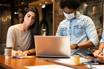 Serious brazilian business colleagues with face mask working and typing in laptop in open office space. Connection, workplace, networking, concept..