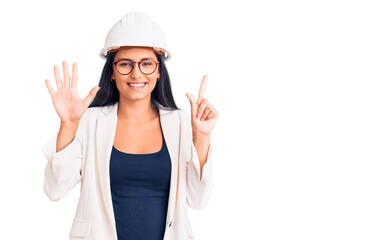 Young beautiful latin girl wearing architect hardhat and glasses showing and pointing up with fingers number seven while smiling confident and happy.