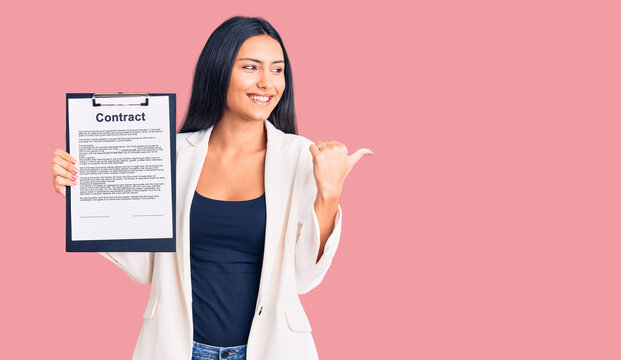 Young beautiful latin girl holding clipboard with contract document pointing thumb up to the side smiling happy with open mouth