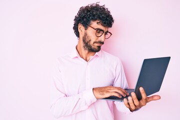 Handsome young man with curly hair and bear working using computer laptop thinking attitude and sober expression looking self confident