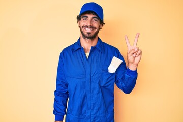 Handsome young man with curly hair and bear wearing builder jumpsuit uniform showing and pointing up with fingers number two while smiling confident and happy.