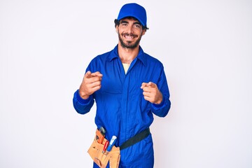 Handsome young man with curly hair and bear weaing handyman uniform pointing fingers to camera with happy and funny face. good energy and vibes.