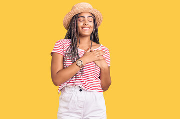 Young african american woman with braids wearing summer hat smiling with hands on chest with closed eyes and grateful gesture on face. health concept.