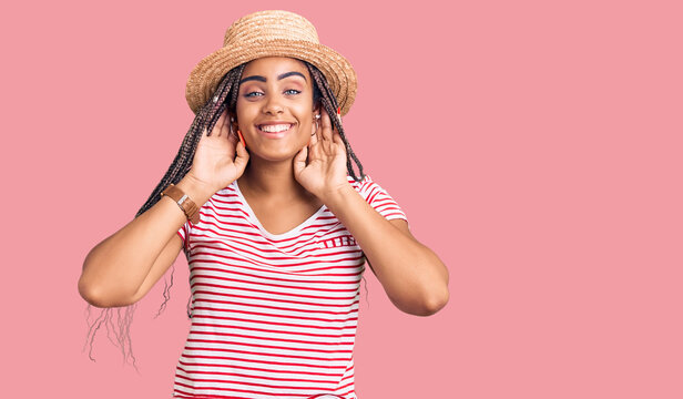 Young african american woman with braids wearing summer hat smiling pulling ears with fingers, funny gesture. audition problem