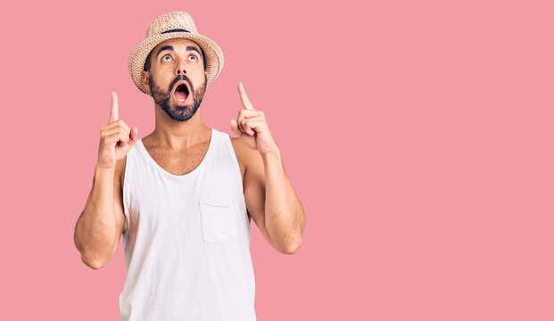 Young hispanic man wearing casual summer hat amazed and surprised looking up and pointing with fingers and raised arms.