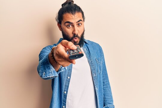 Young arab man holding television remote control scared and amazed with open mouth for surprise, disbelief face