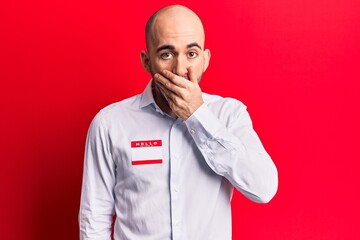 Young handsome bald man wearing hello my name is sticker covering mouth with hand, shocked and afraid for mistake. surprised expression