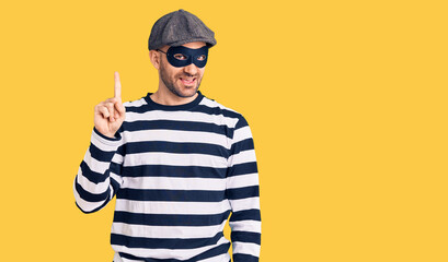 Young handsome man wearing burglar mask pointing finger up with successful idea. exited and happy. number one.