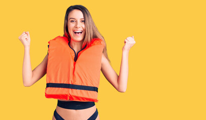 Young beautiful girl wearing life jacket screaming proud, celebrating victory and success very excited with raised arms