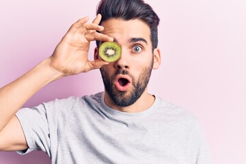 Young handsome man with beard holding kiwi over eye scared and amazed with open mouth for surprise, disbelief face