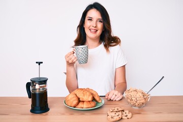 Young beautiful brunette woman sitting on the table having breakfast looking positive and happy standing and smiling with a confident smile showing teeth