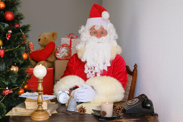adult santa claus in red suit sits at table, Christmas tree is beautifully decorated with balls,...