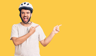Young hispanic man wearing bike helmet smiling and looking at the camera pointing with two hands and fingers to the side.