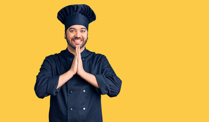 Young hispanic man wearing cooker uniform praying with hands together asking for forgiveness smiling confident.