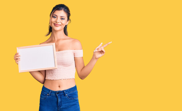 Young beautiful woman holding empty white chalkboard smiling happy pointing with hand and finger to the side