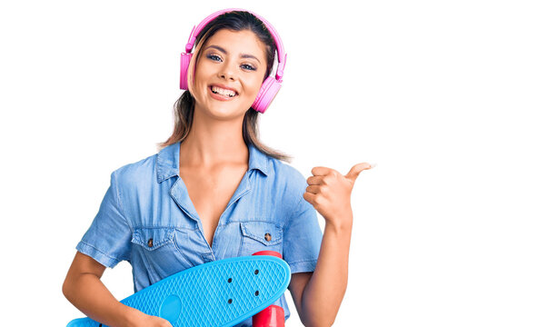 Young beautiful woman wearing headphones and holding skate pointing thumb up to the side smiling happy with open mouth