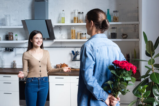 Back View Of Man Holding Bouquet Of Roses Behind Back Near Excited Woman Standing With Open Arms In Kitchen