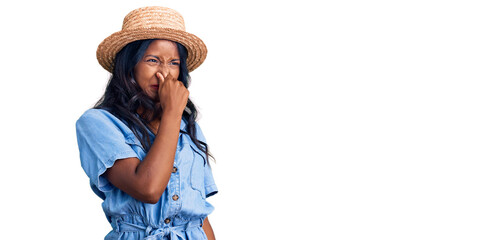 Young indian girl wearing summer hat smelling something stinky and disgusting, intolerable smell,...