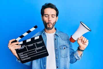 Young hispanic man holding video film clapboard and megaphone making fish face with mouth and...