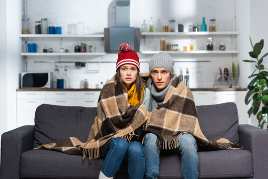 Cold Couple, Wrapped In Plaid Blanket, Wearing Warm Hats, Looking At Camera While Sitting On Couch In Kitchen