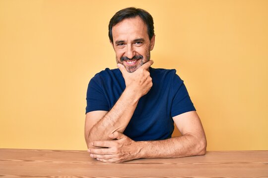 Middle age hispanic man wearing casual clothes sitting on the table looking confident at the camera smiling with crossed arms and hand raised on chin. thinking positive. - Powered by Adobe