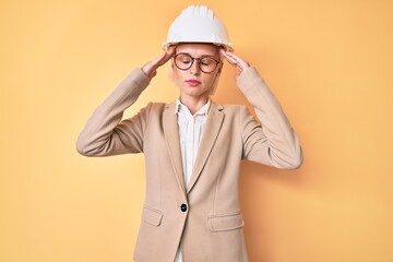 Young blonde woman wearing architect hardhat with hand on head, headache because stress. suffering migraine.