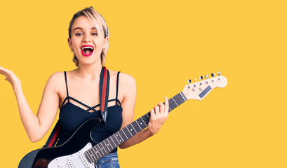 Fototapeta premium Young beautiful blonde woman playing electric guitar celebrating victory with happy smile and winner expression with raised hands