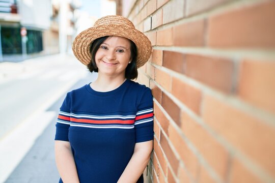 Beautiful brunette woman with down syndrome wearing a summer hat at the town on a sunny day leaning on a bricks wall