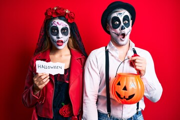 Couple wearing day of the dead costume holding pumpking and halloween paper in shock face, looking skeptical and sarcastic, surprised with open mouth