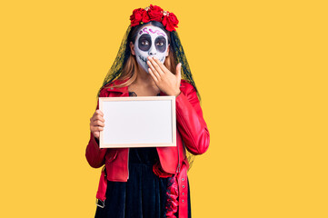 Woman wearing day of the dead costume holding empty white chalkboard covering mouth with hand, shocked and afraid for mistake. surprised expression