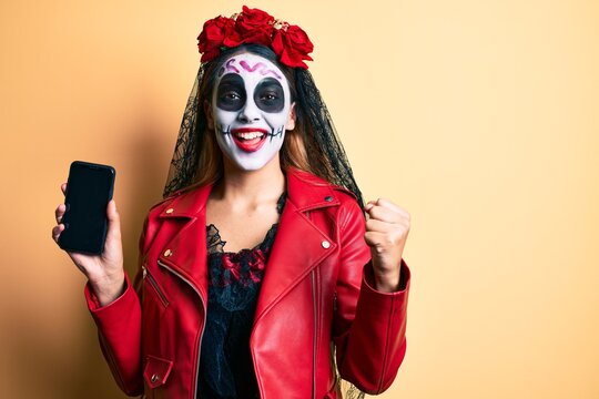 Woman Wearing Day Of The Dead Costume Holding Smartphone Showing Screen Screaming Proud, Celebrating Victory And Success Very Excited With Raised Arms