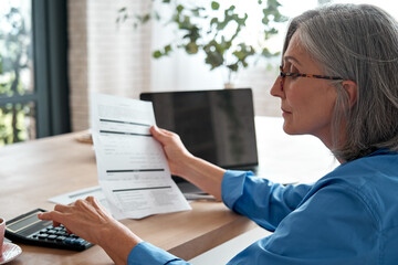 Senior mature business woman holding paper bill using calculator, old lady managing account finances, calculating money budget tax, planning banking loan debt pension payment sit at home office table. - obrazy, fototapety, plakaty