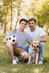 Obraz premium father sitting on grass near teenager son with football and golden retriever