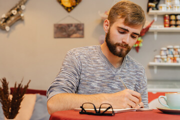 A bearded man sits at a table in a cafe and writes in a notebook.