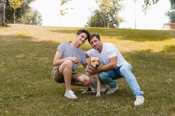 father hugging teenager boy near golden retriever while sitting on grass in park