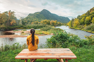 Fototapeta premium Camping nature woman sitting at picnic table enjoying view of wilderness river in Quebec and autumn foliage forest, Canada travel. Parc de la Jacques-Cartier, Quebec.