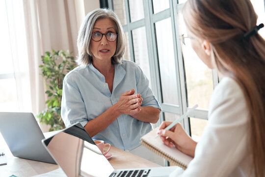 Mature Old Female Mentor Or Manager Training Young Intern Explaining Sharing Professional Skills In Office. Middle Aged Senior Business Woman Teacher Speaking To Student Teaching Trainee At Meeting.