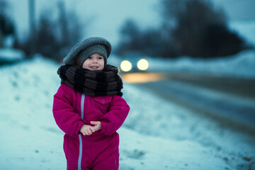 Fototapeta na wymiar .A little girl stands in the snow by the road in the evening