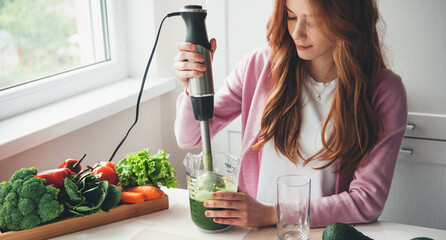 Caucasian ginger woman with freckles is using an electrical squeezer for making fresh green vegetable juice at home