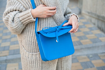 Young fashionable woman wearing gray knit cardigan. She is holding blue leather handbag in hands....