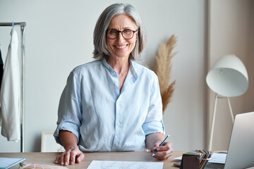 Happy middle aged stylish woman fashion designer wears glasses standing at workplace desk,...