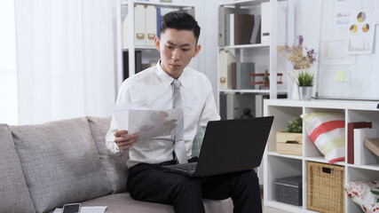 asian korean male employee is reading financial analysis on paper and typing up data on laptop computer while sitting on sofa in living room.