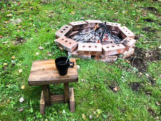 A cup of coffee stands on a stool on the grass near a fire surrounded by bricks. Vacation in the...