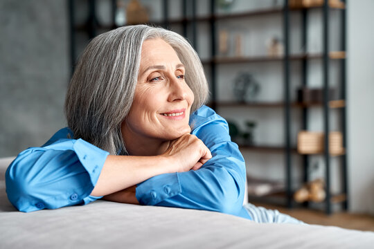 Happy relaxed mature old woman resting dreaming sitting on couch at home. Smiling mid aged woman relaxing. Peaceful serene grey-haired lady feeling peace of mind enjoying lounge on sofa and thinking.