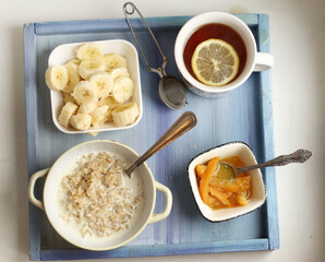 healthy breakfast on blue tray - porridge, cut banana, orange jam, lemon tea with spoon and tea strainer