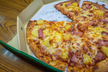 Close up hot fresh-baked Hawaiian pizza cheese melted in carton box on the table at the home background. Enjoying the menu takes away junk food. Fast food takeout delivery.